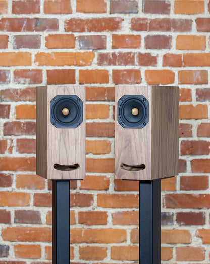 Closer Acoustics OGY bookshelf loudspeakers – pair in walnut veneer, side profile view against a brick wall, emphasizing their stylish design in a modern interior